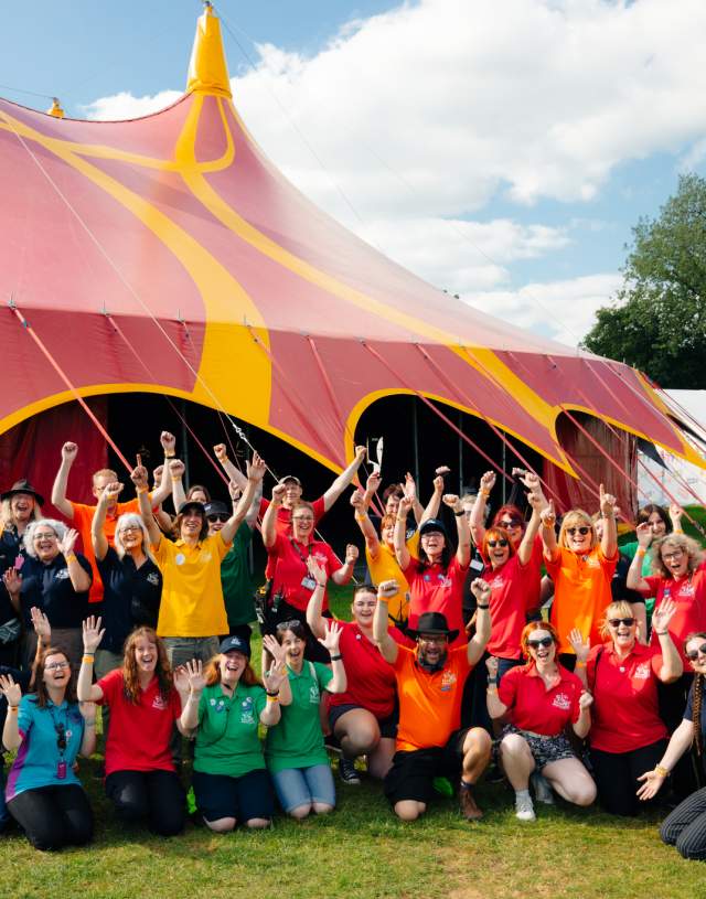 The team from Big Malarkey Festival in colourful t shirts in a group in front of a big red and yellow marquee.