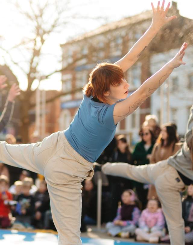 Three dancers stand on one leg in Trinity Square with a crowd surrounding them