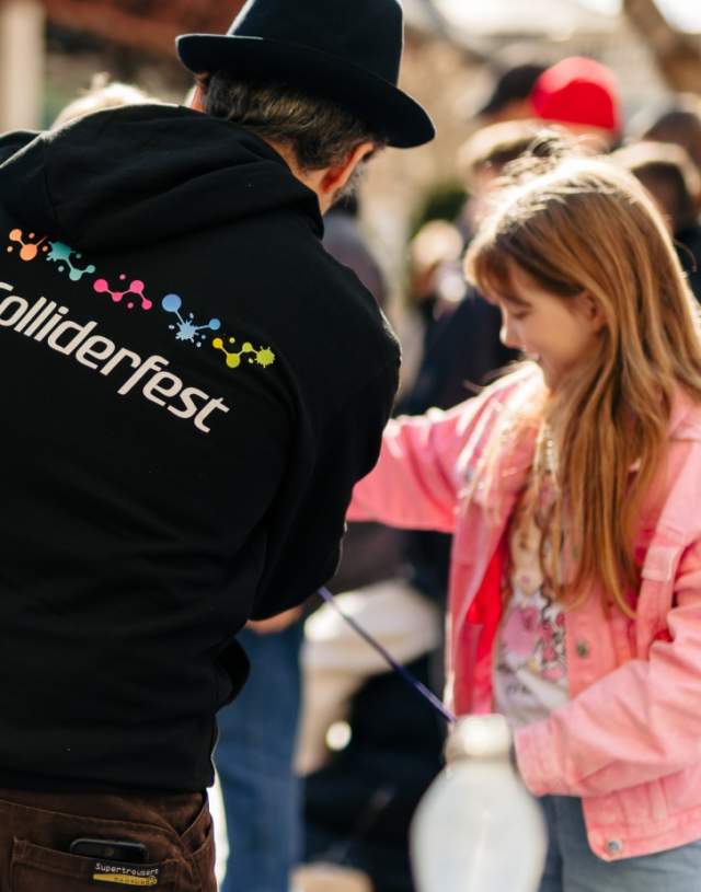 Man in a black Colliderfest hoody shows a science trick to a young girl