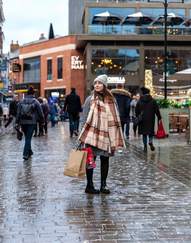 A woman in a scarf is stood in the centre of the Cornhill Quarter in Lincoln, holding shopping bags.