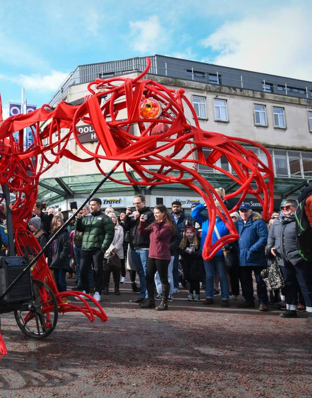 a large moving dragon puppet being controlled by a puppeteer in a parade with crowd watching on