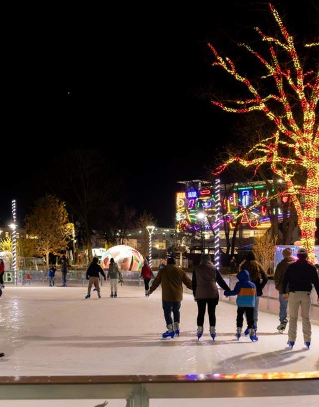 Ice skating rink illuminated at night, surrounded by festive lights and trees. People skate and enjoy the vibrant atmosphere.