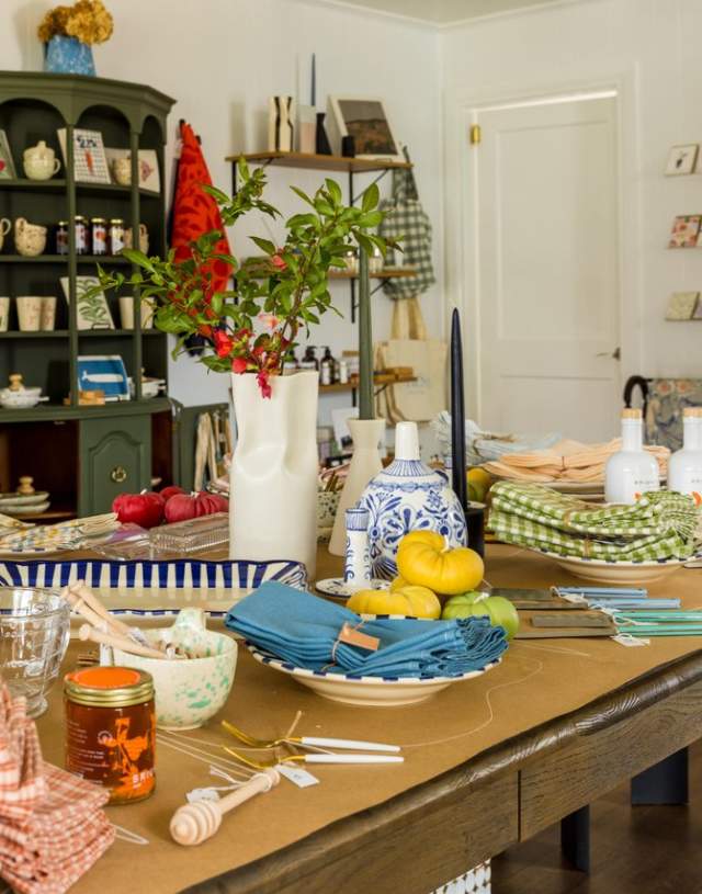 A cozy shop interior featuring a wooden table set with colorful tableware, candles, and fresh produce. Shelves display various home goods and decor.