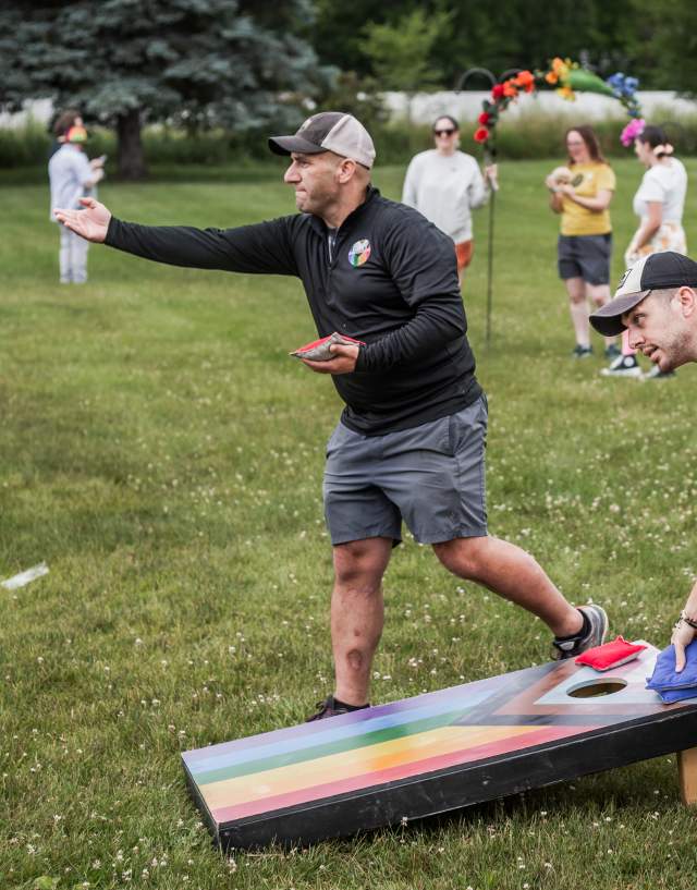 Two men playing cornhole at a Pride event in Mason. There are rainbow colors on the cornhole board and people in the background.