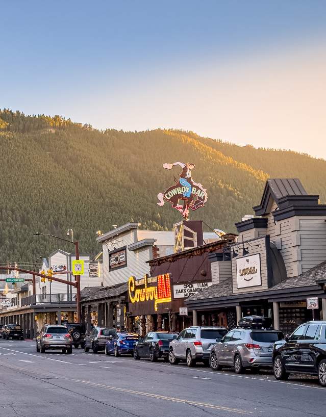 A western-style downtown street lined with shops and parked cars sits beneath forested mountains, with a large “Cowboy Bar” sign visible above the buildings in warm evening light.