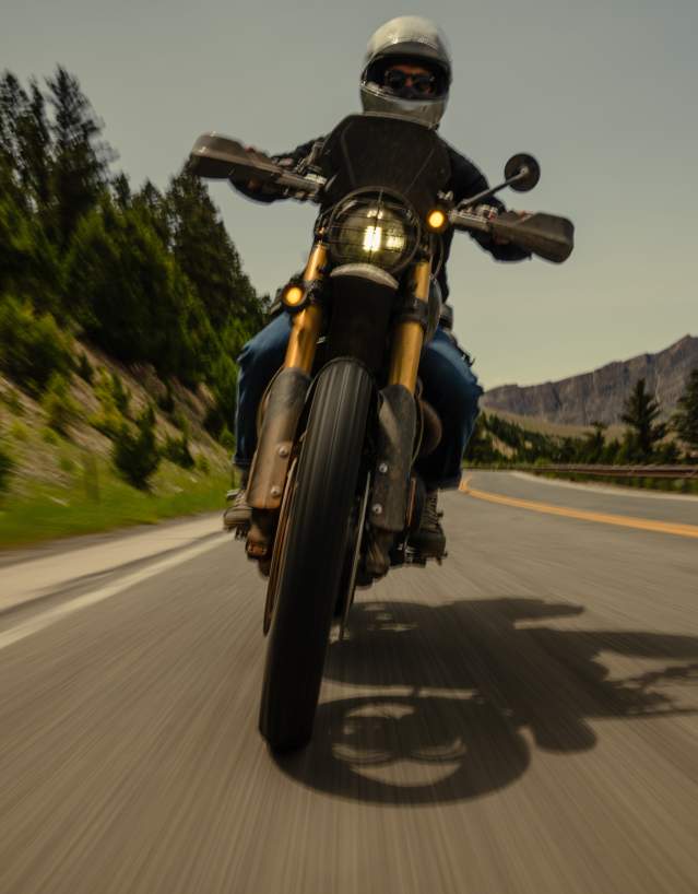 A motorcyclist rides along a winding mountain road, with forested hills and rocky cliffs in the background under a clear sky.