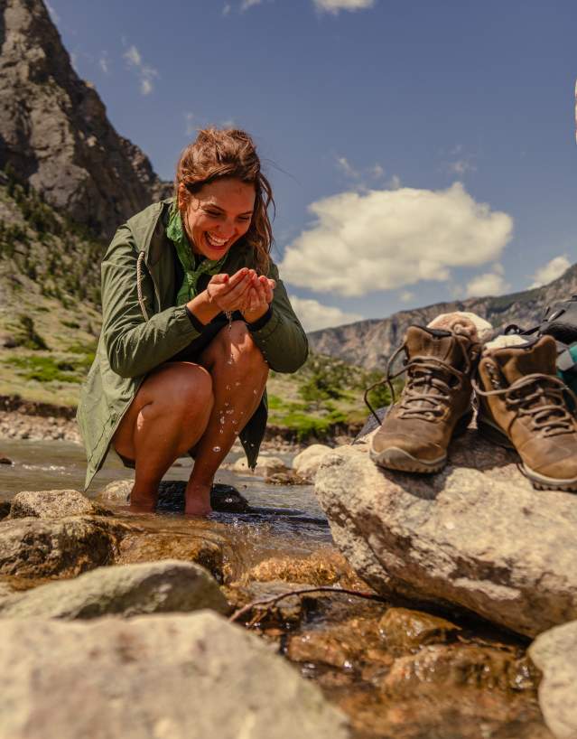 A woman kneels in a shallow mountain stream, smiling as she cups water in her hands, while hiking boots and a backpack rest on nearby rocks with forested cliffs in the background.