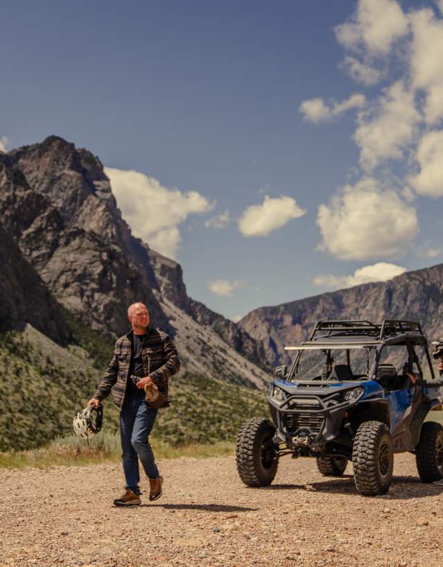 Two people stand beside an off-road utility vehicle on a gravel road in a mountain valley, with steep rocky cliffs and scattered clouds in the sky behind them.