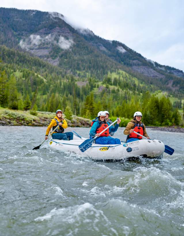 Three people paddle an inflatable raft through a flowing river, wearing helmets and life jackets, with forested mountains rising in the background.