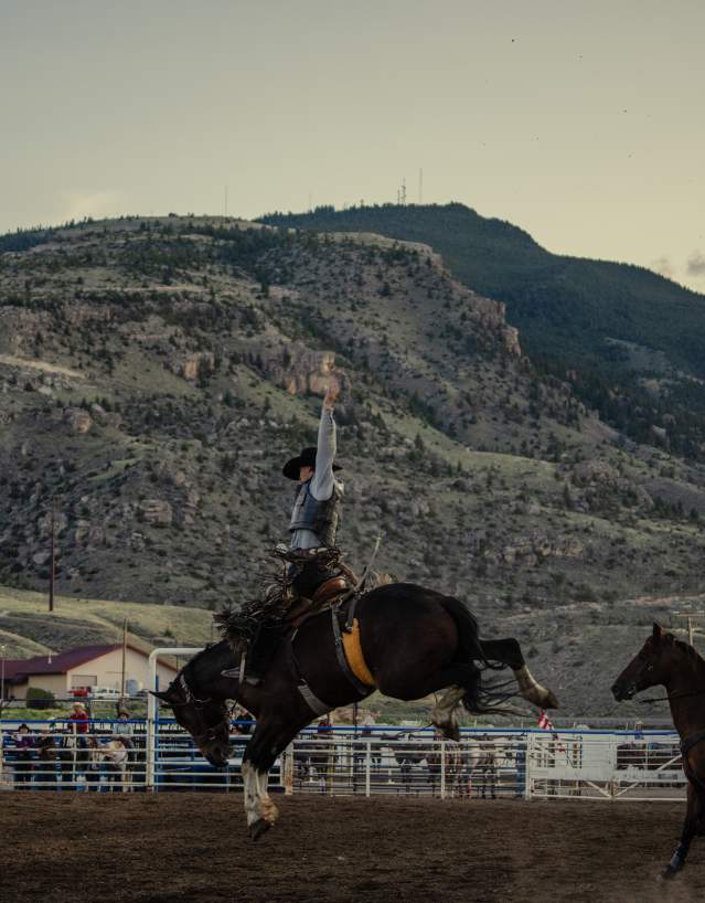 A rodeo rider raises one arm while riding a bucking horse inside an outdoor arena, with another mounted rider nearby and tree-covered mountains rising in the background at sunset.