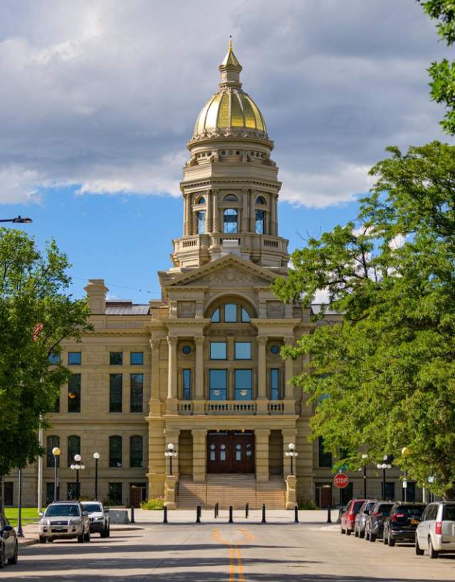 A street lined with parked cars and rows of trees, and in the background, the Wyoming State Capitol.