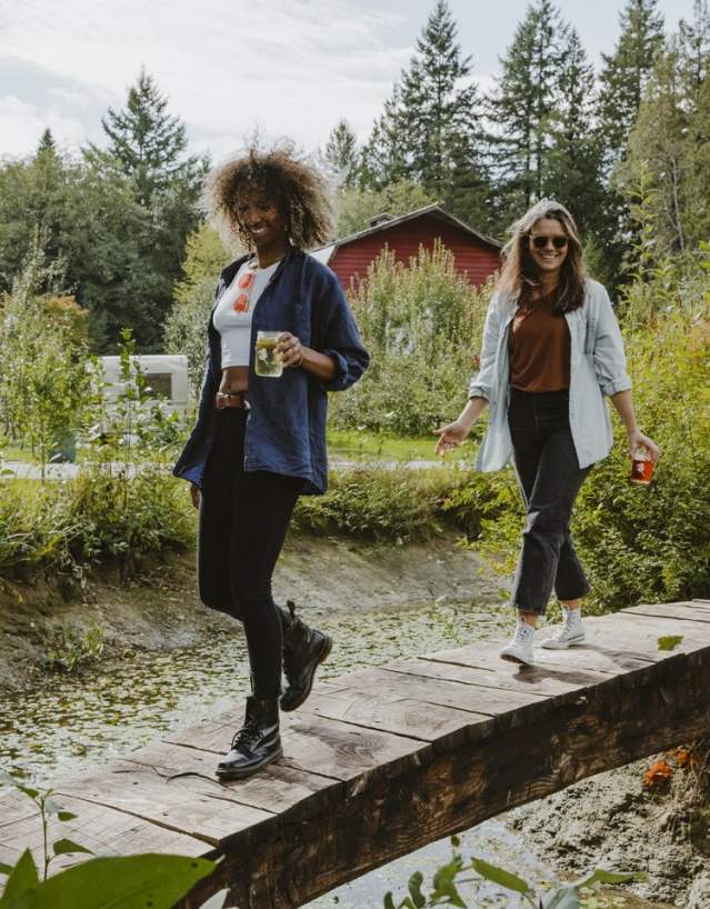 Two women walk across a small bridge, carrying glasses of cider.