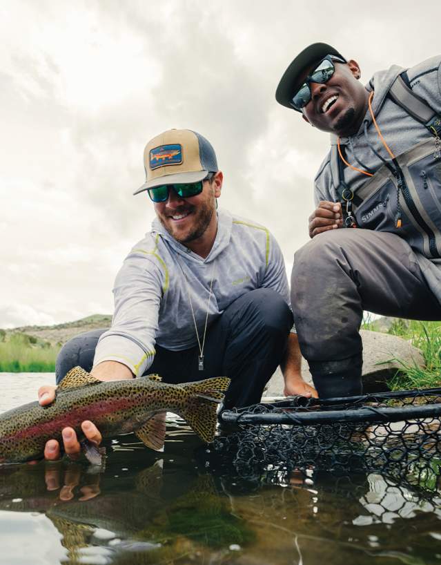 Two people kneel at the edge of a river, smiling as they hold a freshly caught fish above the water, with fishing gear and grassy riverbanks visible around them.