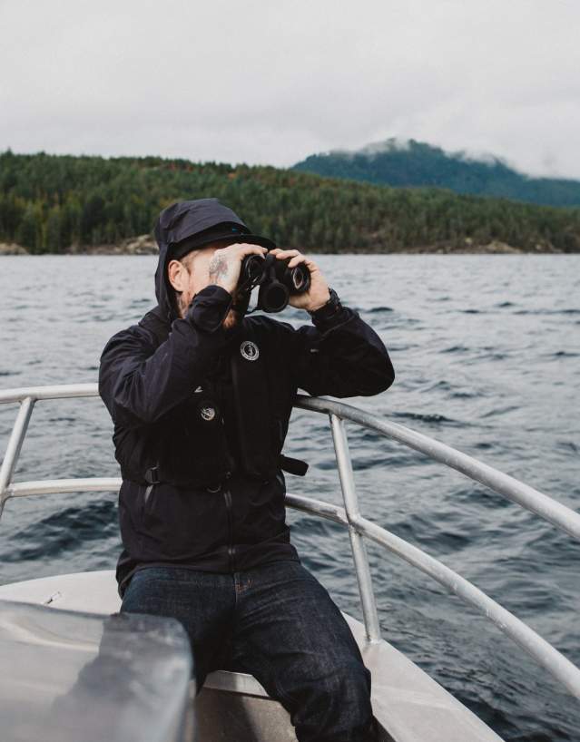 A man sits on a fishing boat with Nelson Island in the background, looking through binoculars.