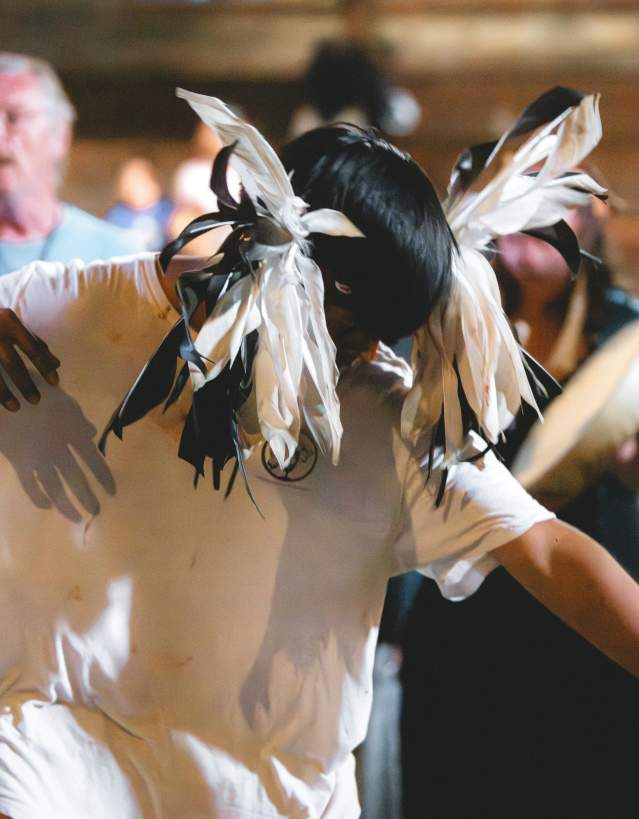 A dancer wearing a black and white feathered headdress performs inside a gathering space while people watch and drums play in the background.