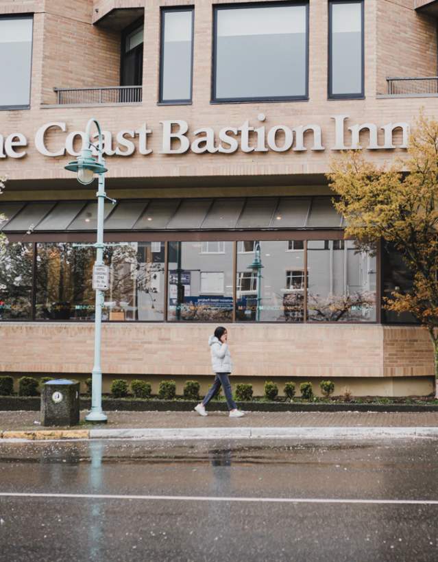A person walks past The Coast Bastion Inn on a rainy day, with wet pavement and trees lining the street.
