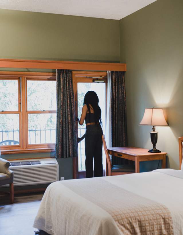 A cozy hotel room with a bed, desk, and seating area as a person opens a balcony door to let in natural light.