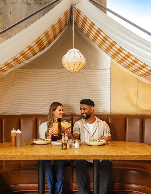 Young Couple sitting and holding drinks in a booth with a tent canopy overhead, at Union Rec