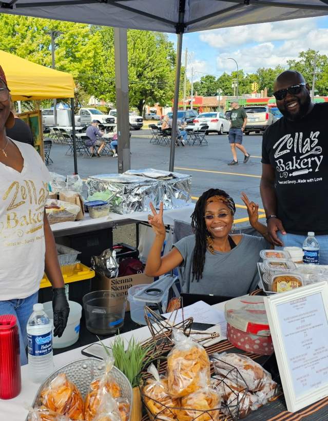Three people pose behind a taple filled with baked goods for sale, set up in a parking lot. A person's T-shirt reads Zella's Bakery