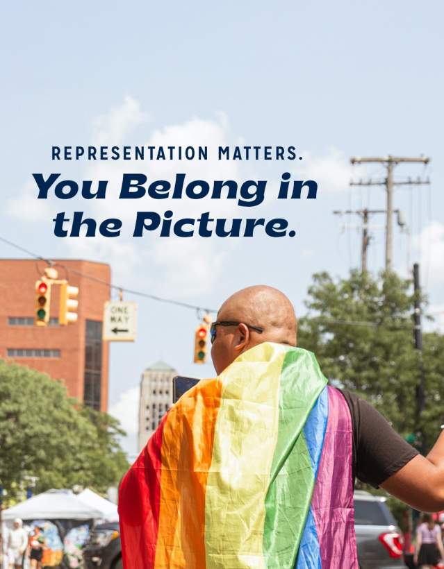 Person with back towards camera, wearing rainbow flag as a cape, waving pride flags in their right hand as they walk away. The photo has text that has been added: "Representation Matters. You belong in the picture."