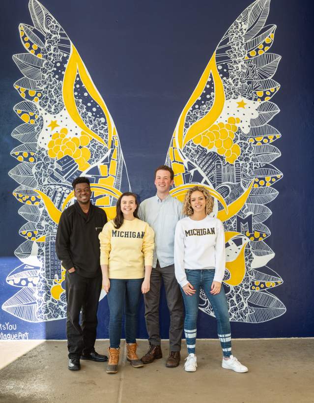 Four U-M students in front of blue & maize wings mural