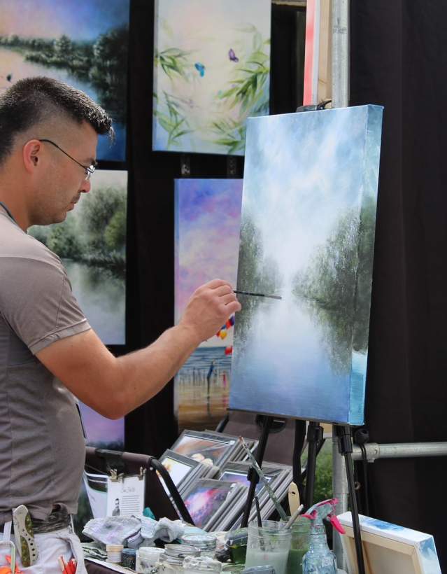 Man painting a blue and green landscape painting at an outdoor fair