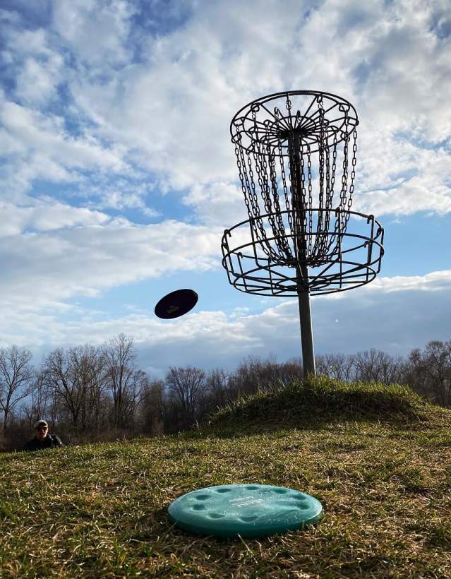 Person in background throwing a disc golf frisbee at goal in foreground