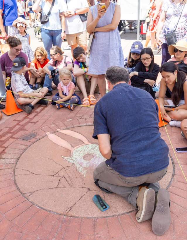 Man with back to camera drawing with chalk on pavement while crowd looks on