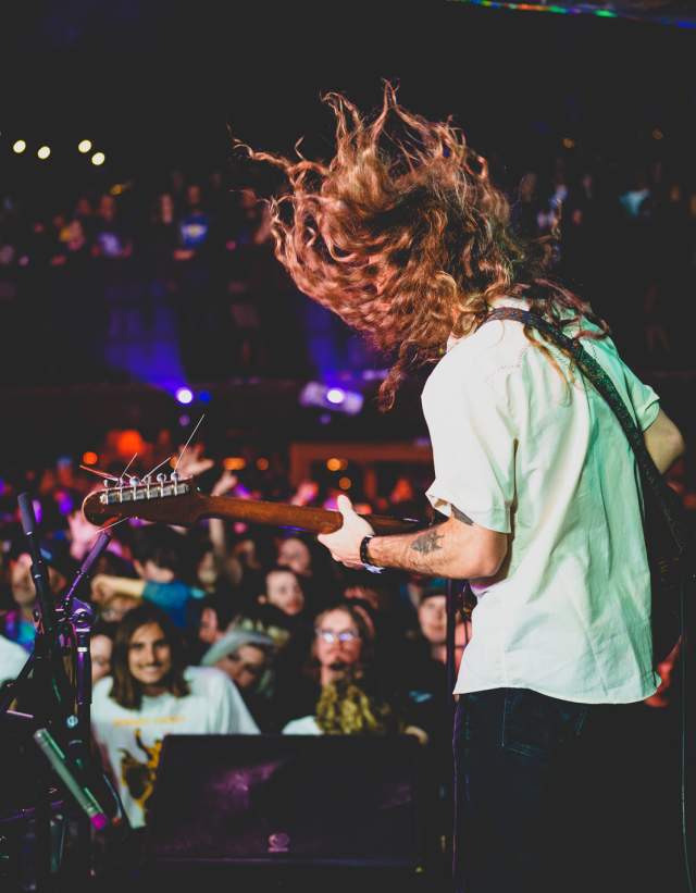 Guitar player headbanging with hair in motion while a large crowd is smiling and dancing.