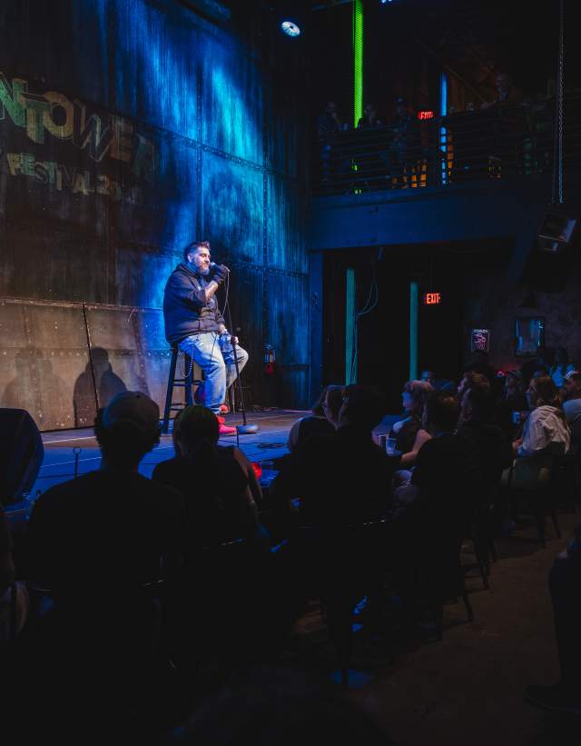 A man sitting on a stool onstage with a mic during a comedy set at Vulcan Gas Company.