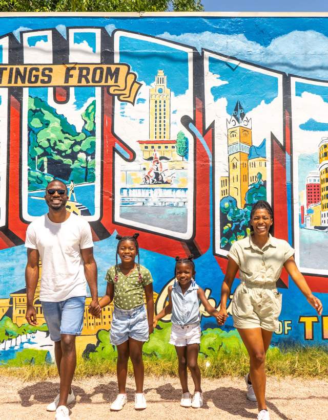 Family of four with two young daughters hold hands and smile at camera in front of postcard mural that reads Greetings from Austin Texas