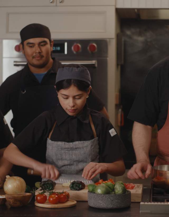 Four student chefs standing in a designed commercial kitchen, three of the students watching one of them chopping a vegetable.