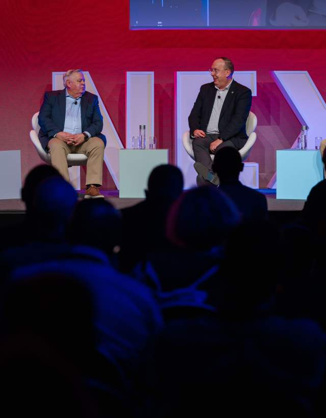 A woman sitting on a stage with three men to her right, speaking on a panel in front of a dark room of attendees.