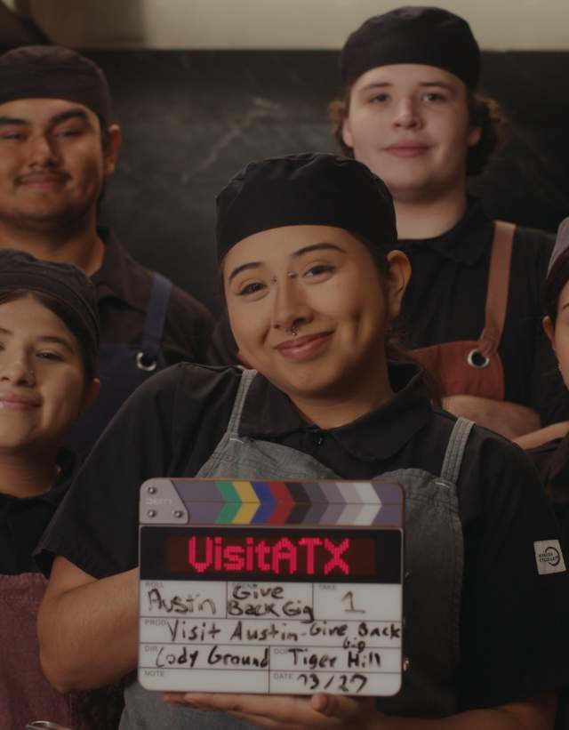 Five student chefs in aprons standing in a commercial kitchen, holding a clapper board with details from the Give Back Gig promo video.