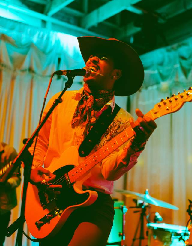 Musician in a cowboy hat singing into a mic while playing guitar with colorful lights surrounding him.