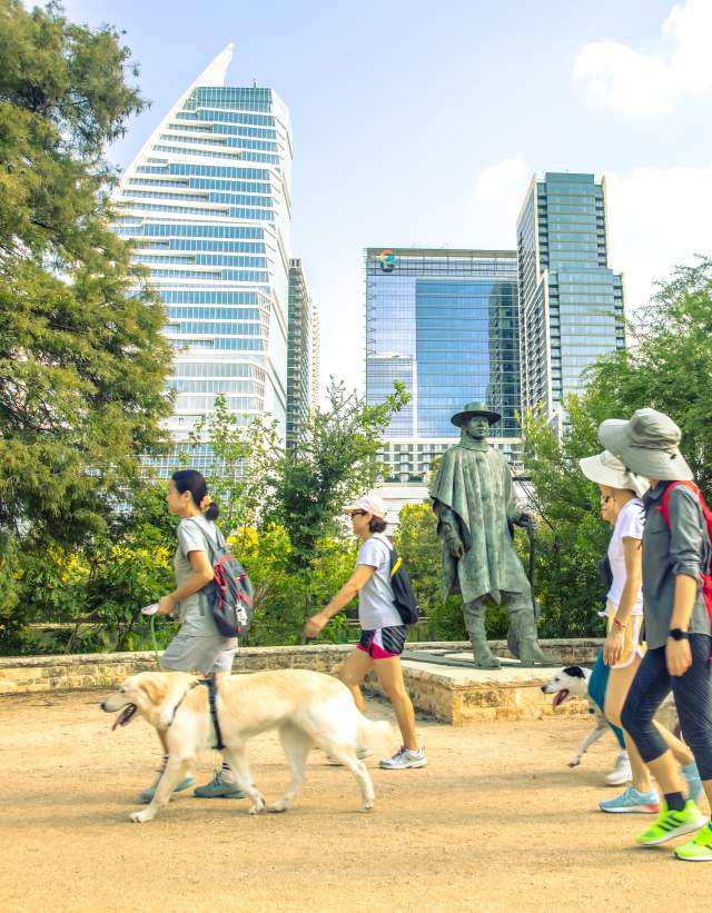 Men and women walking past the Stevie Ray Vaughan statue on the Butler Hike and Bike Trail with their dogs.