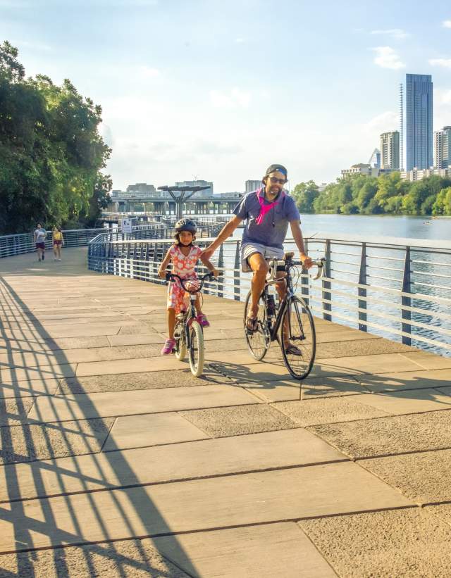 Image of a man and daughter riding bikes on the Butler Hike and Bike trail boardwalk with Austin Downtown in the background.