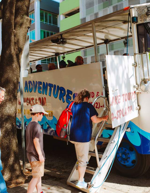 A woman, man and child loading on to the Austin Duck Adventure tour bus, outside of the Austin Visitor Center.