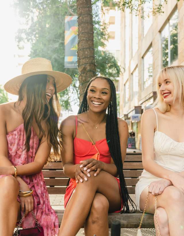 One Asian woman, one Black woman and one white woman sit on a sidewalk bench in front of shops