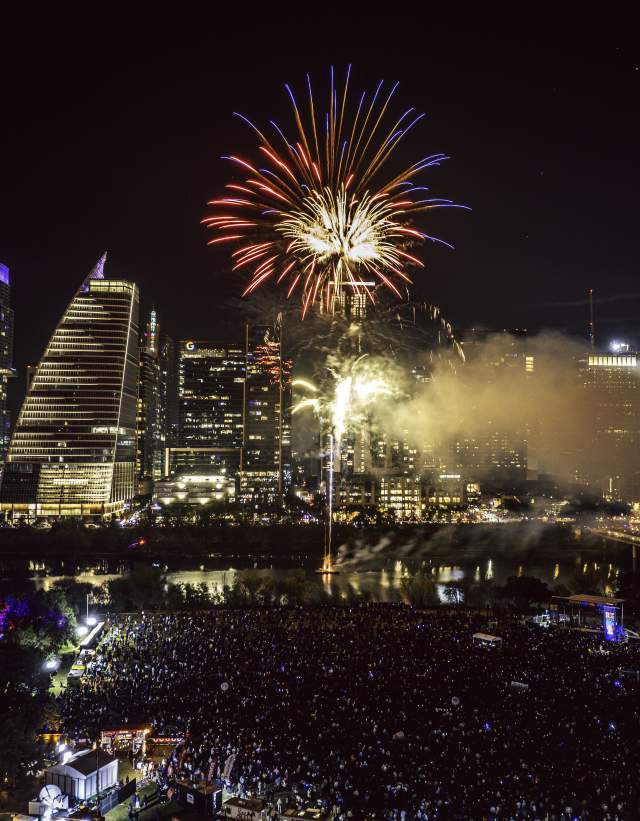 Red fireworks explodes over downtown Austin skyline and Lady Bird Lake at night. Aerial view shows a music stage and crowd gathered below at Auditorium shores.