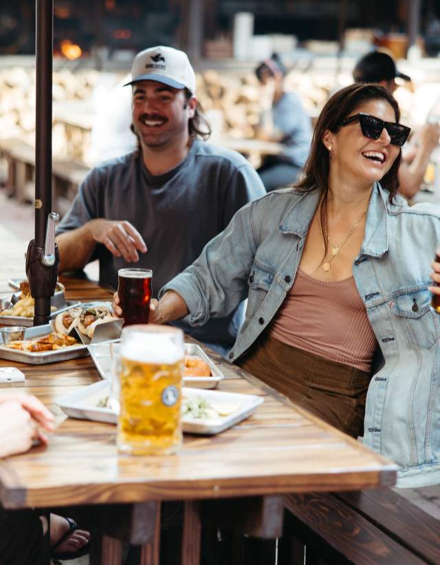 Group of friends sitting, laughing together at a picnic table with steins of beer on a sunny day.