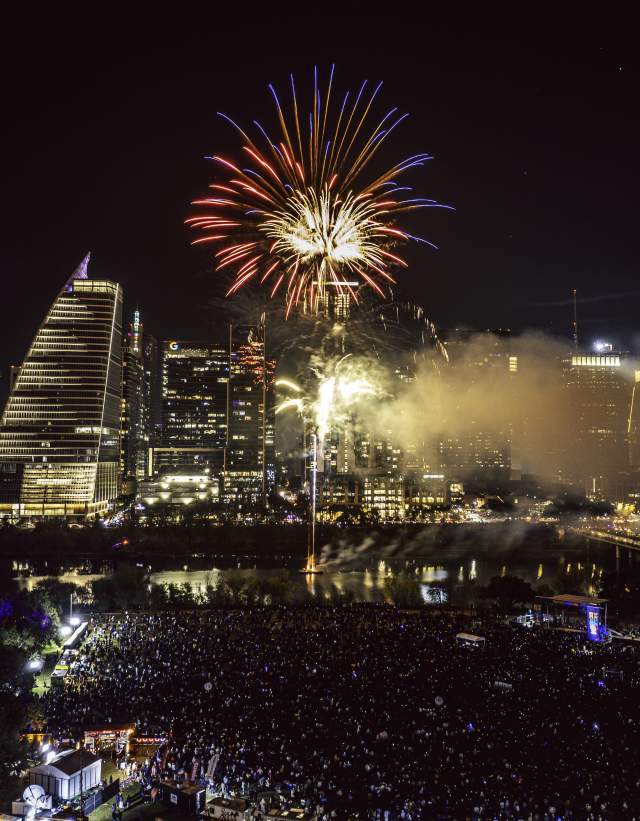 Red fireworks explodes over downtown Austin skyline and Lady Bird Lake at night. Aerial view shows a music stage and crowd gathered below at Auditorium shores.
