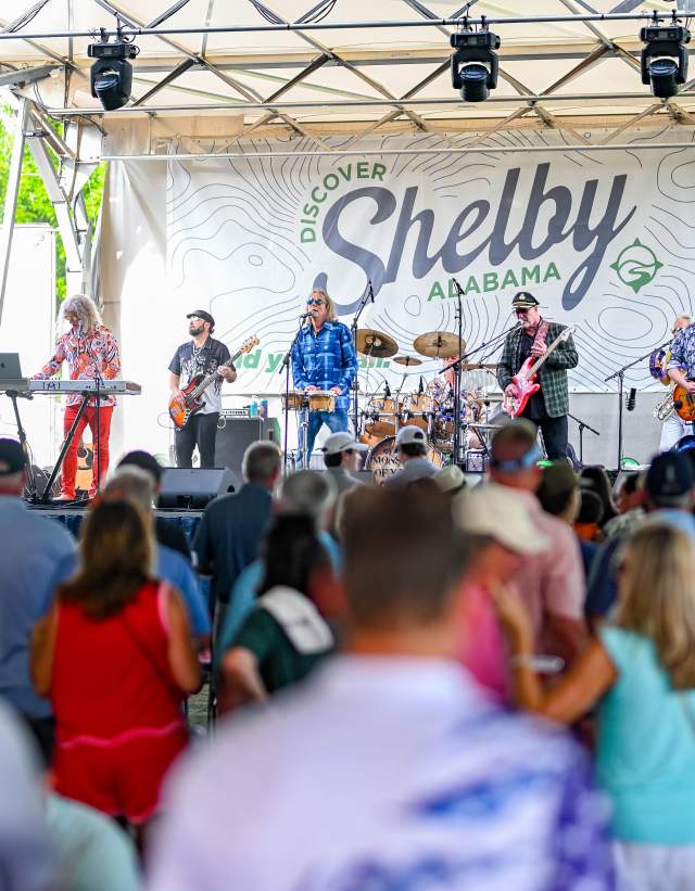 A crowd listens to a band play on a stage featuring a Discover Shelby banner.