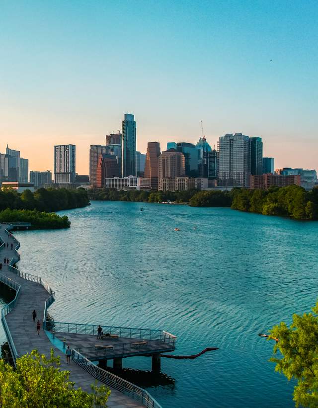 People walking along suspended board walk that follows the shape of the Colorado River, leading to Downtown Austin skyline.