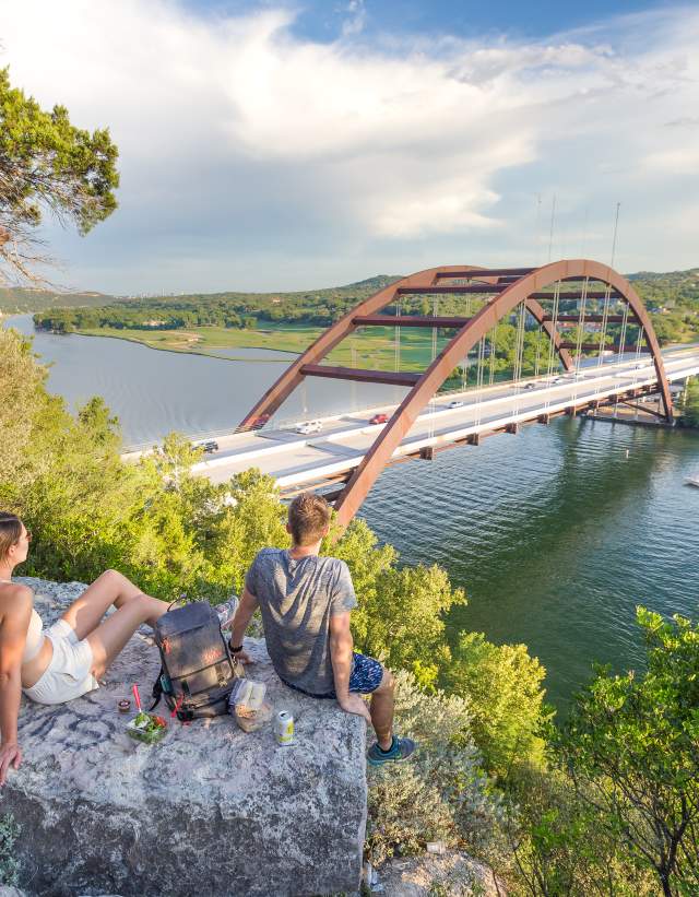 Hikers sitting on a hilltop rock overlooking the Pennybacker Bridge at Lake Austin.