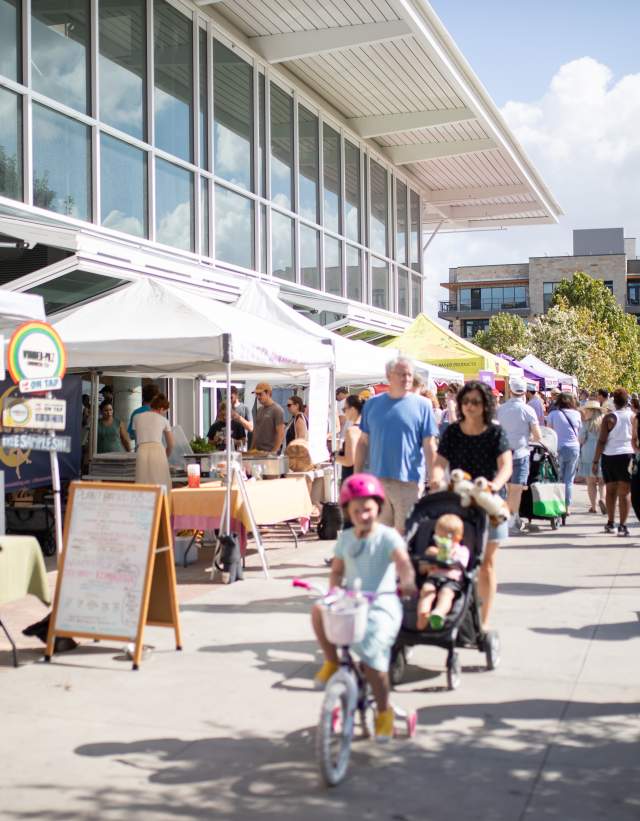 Image of the Texas Farmers Market exterior area of vendors during a market day with people and kids walking around.