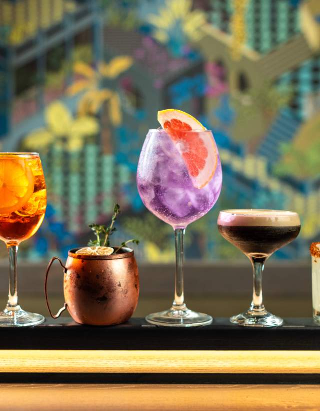 Four colorful cocktails in different shaped glasses sitting on top of a shuffleboard table.