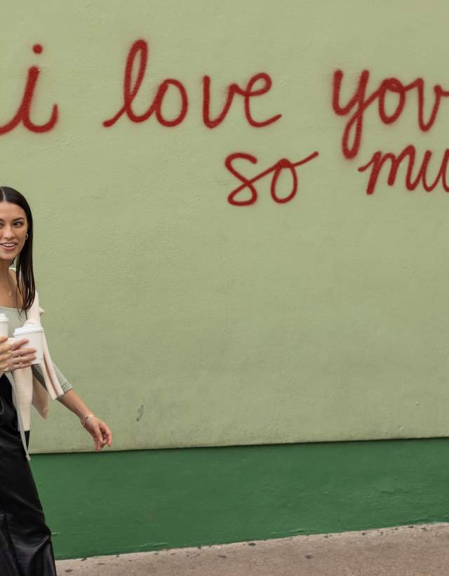 Two women walking in front of the I Love You So Much mural with coffee in hand.