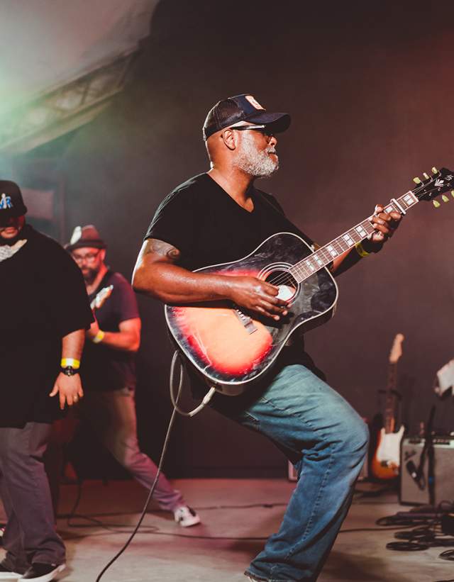 Musician Ray Prim plays an acoustic sunburst guitar on stage at Stubb's in Austin Texas. Two other male musicians stand behind him