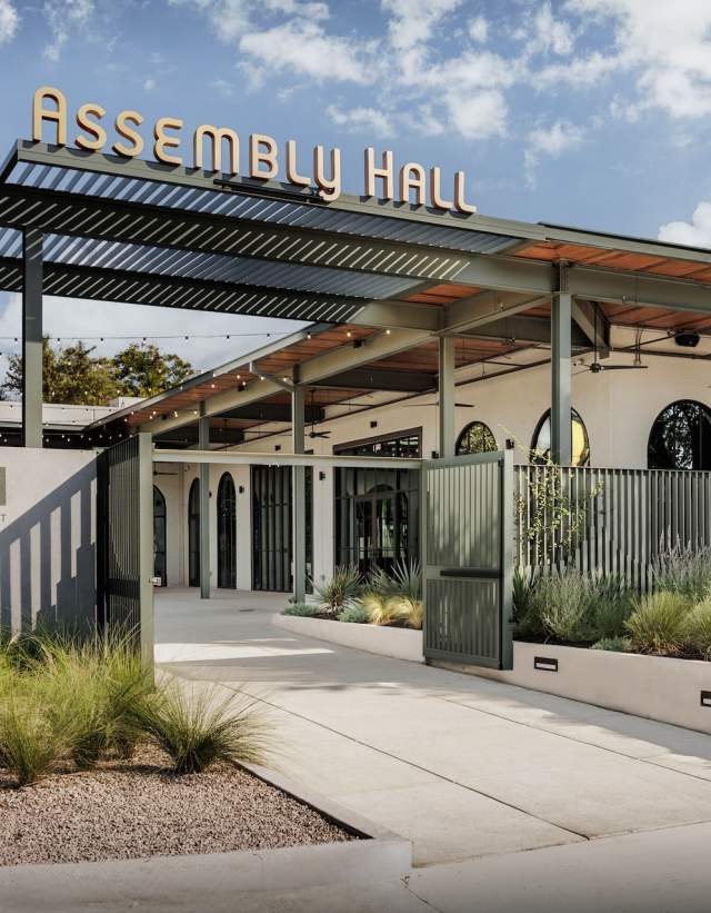 Exterior of an event venue with sign on the roof reading Assembly Hall. The building has a modern facade with dark green fencing, a large patio and native Texas landscaping